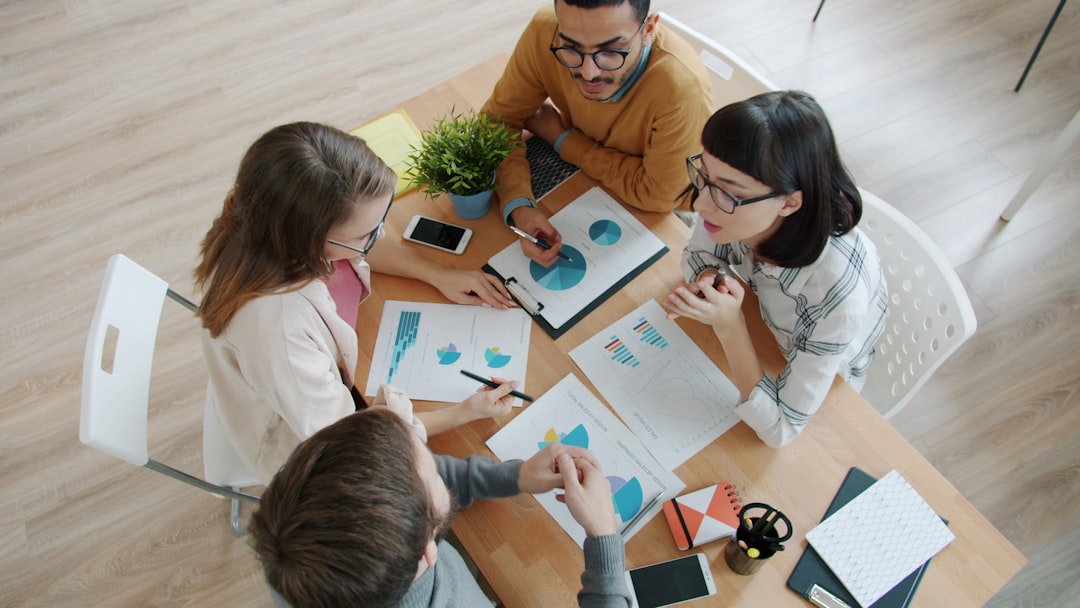 High angle view of young people colleagues discussing project looking at charts sitting at desks in office. Brainstorming, coworking and teamwork concept.